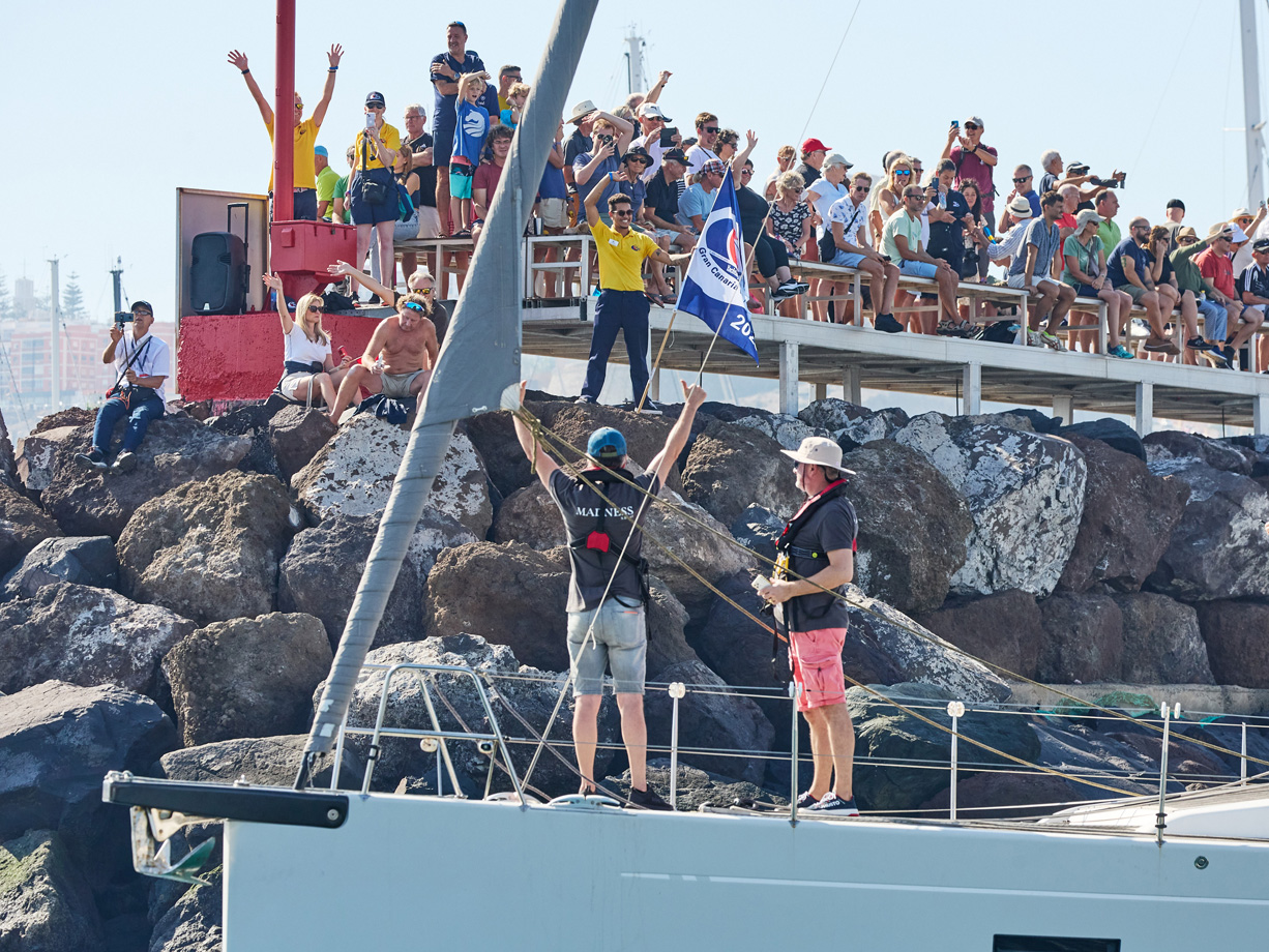 The crew of yacht Madness wave to the hundreds of people lining the sea wall in Las Palmas Marina