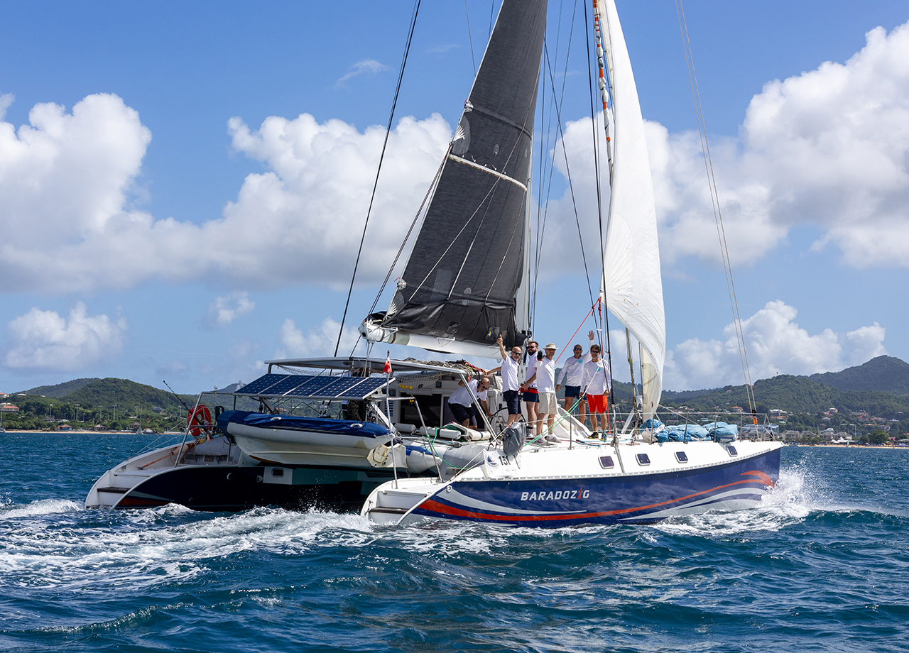 The dark blue catamaran Baradozig sails into Rodney Bay with all crew on deck waving