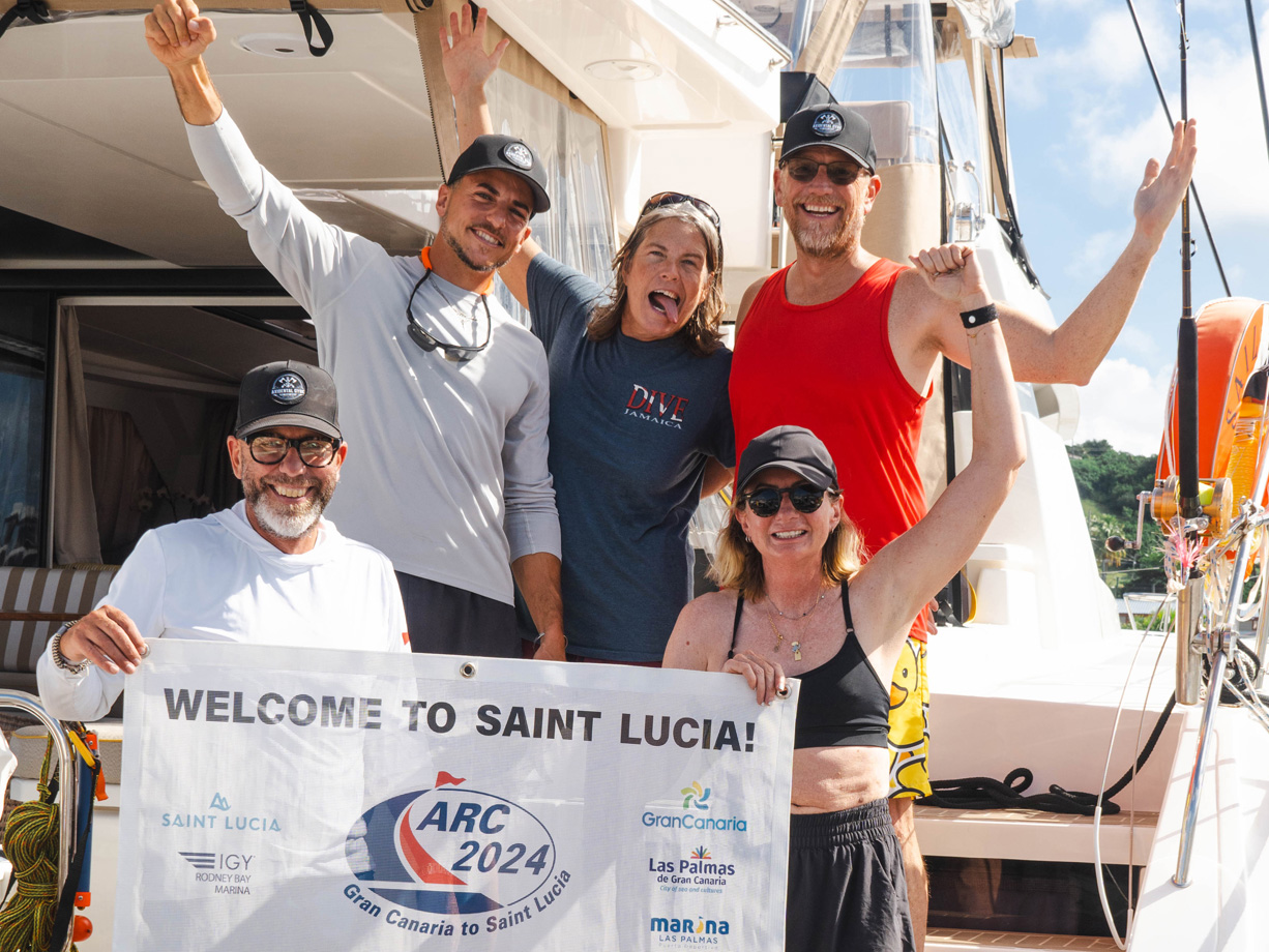 Men and women of the crew of yacht Akula smile and wave on arriving in Saint Lucia in ARC 2024