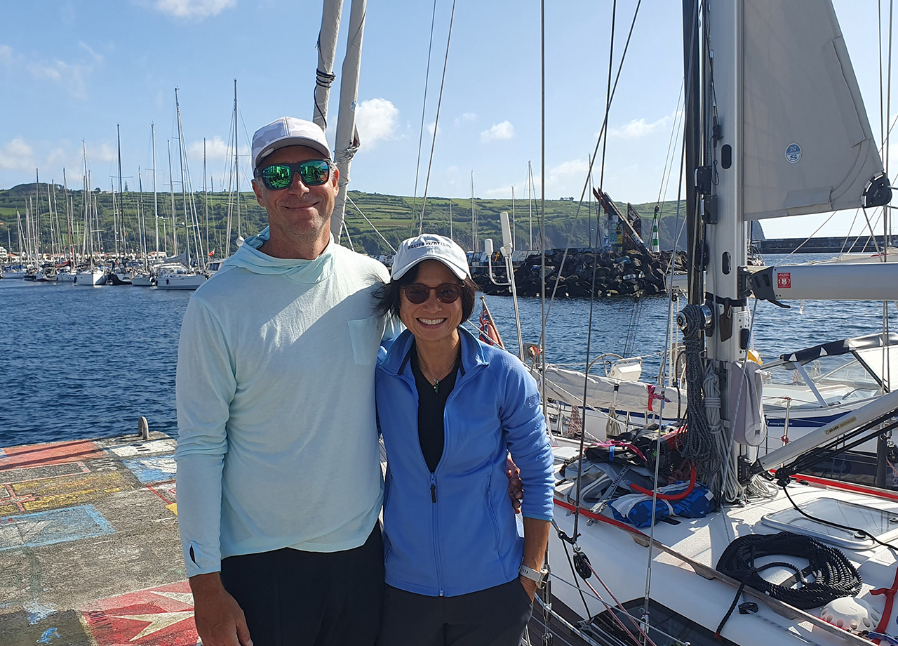 A man and woman stand on the Horta harbour wall in from of yacht Aria Legra in ARC Europe 2025