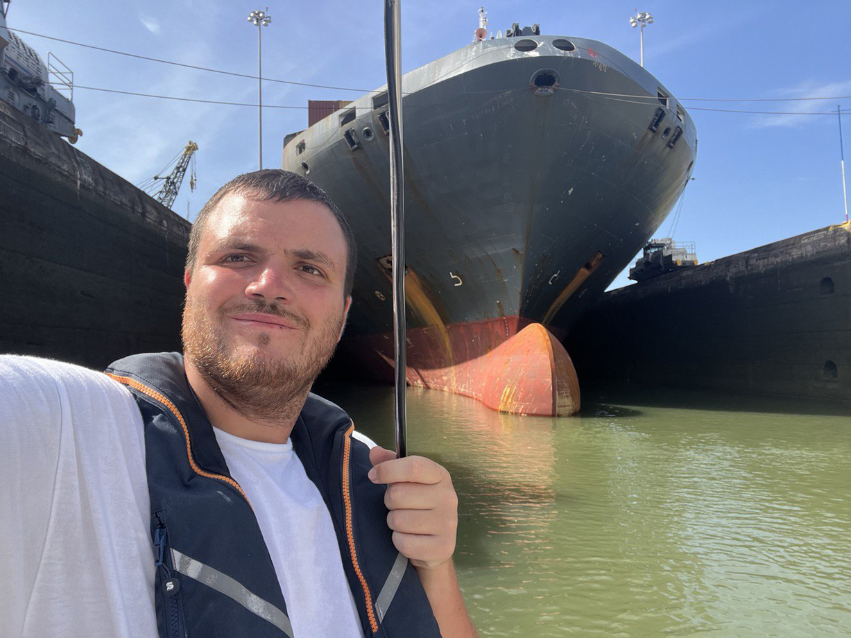 A man poses for a photo onboard a yacht in a lock in the Panama Canal. In the background is the bow of a huge ship