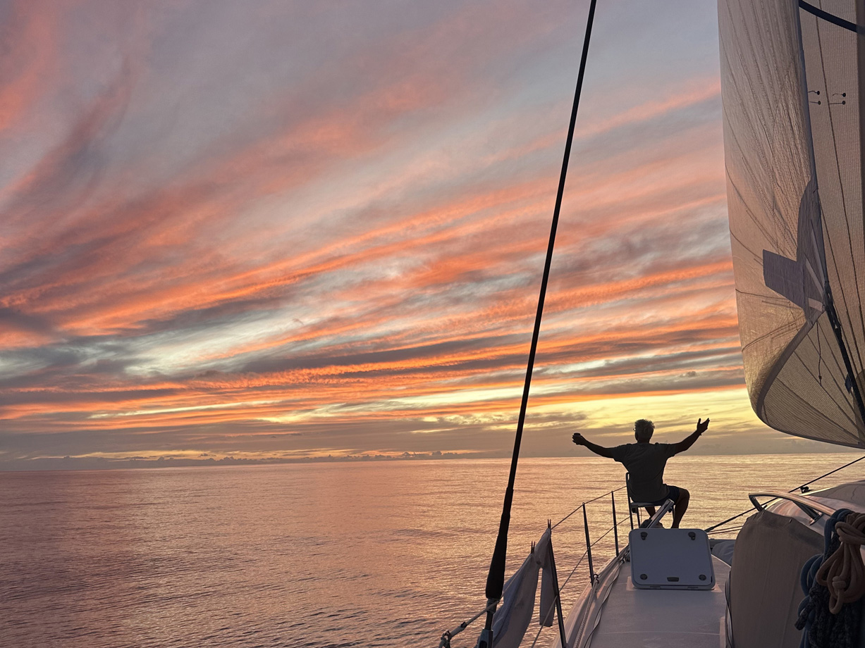 Clouds are lit from below in orange, red and yellow as the sun sets viewed from a boat at sea. A crew member watches with arms outstretched