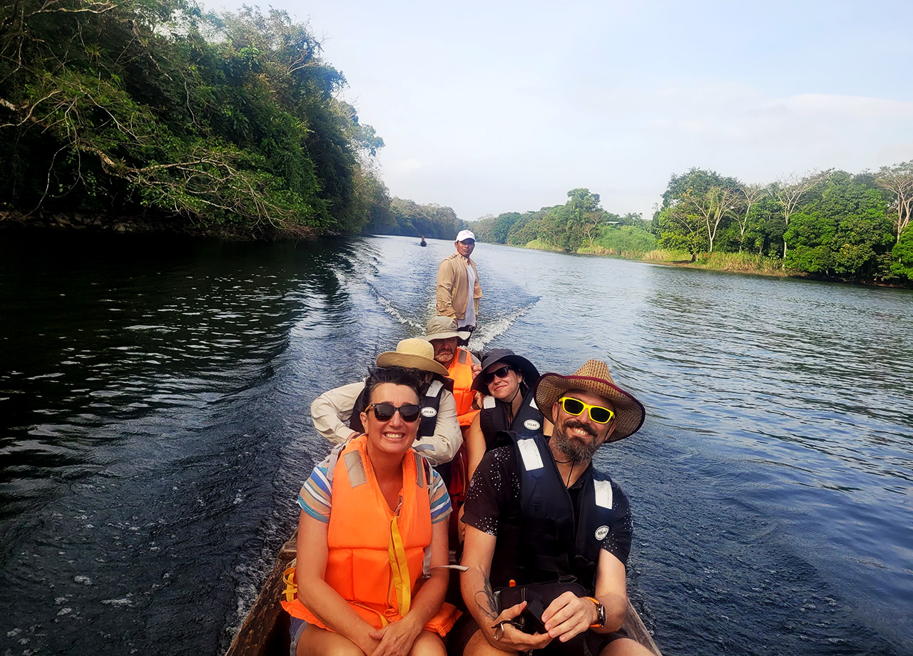 Men and women wearing lifejackets sit in a longboat on a river surrounded by forest in Panama