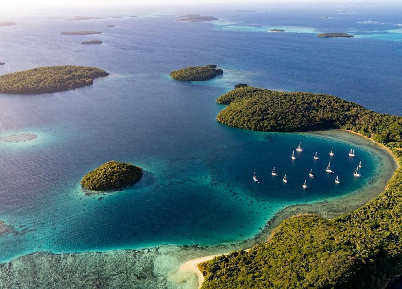 Aeial view of green wooded islands dotted in a blue sea, with a group of yachts anchored in a bay with a white sand beach