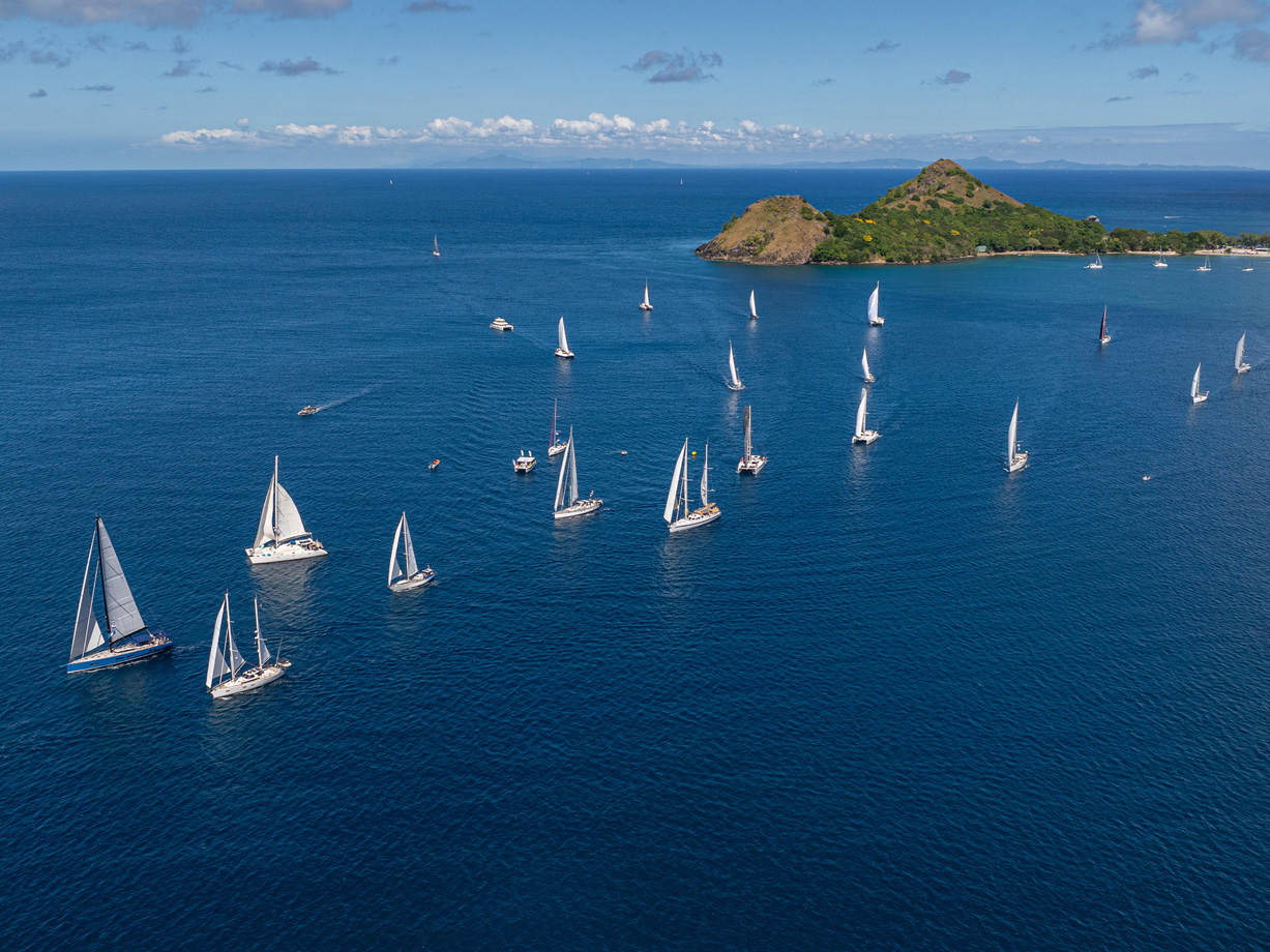 An aerial view of a fleet of yachts with white sails on a calm blue sea with Pigeon Island in the background