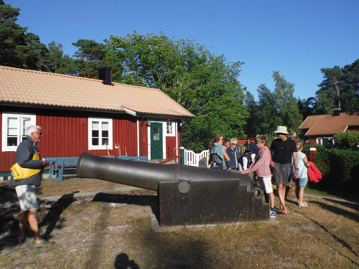 A group of people look at an old cannon next to a red wooden building with white winds