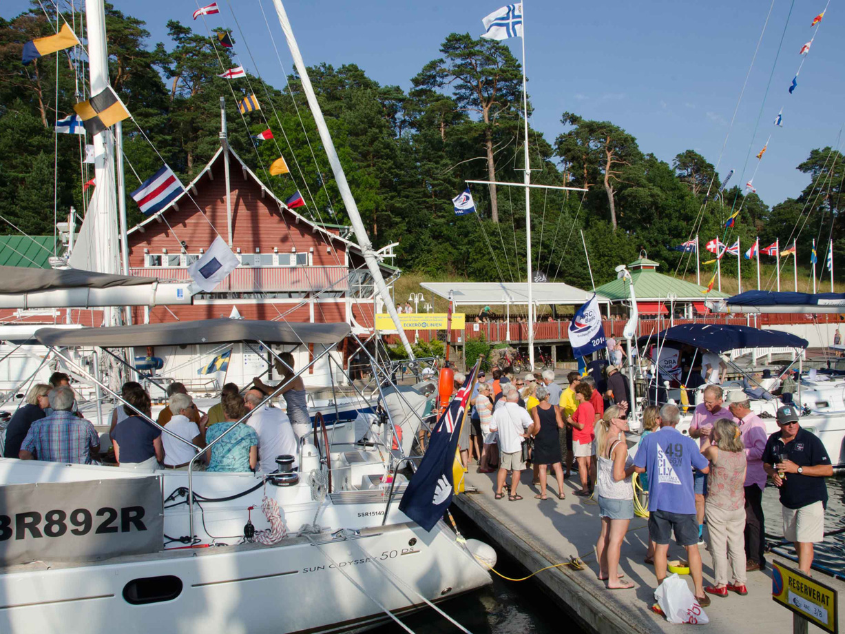A large group of people having a party on a sunny evening on a dock with yachts moored up. Red wooden buildings in the background