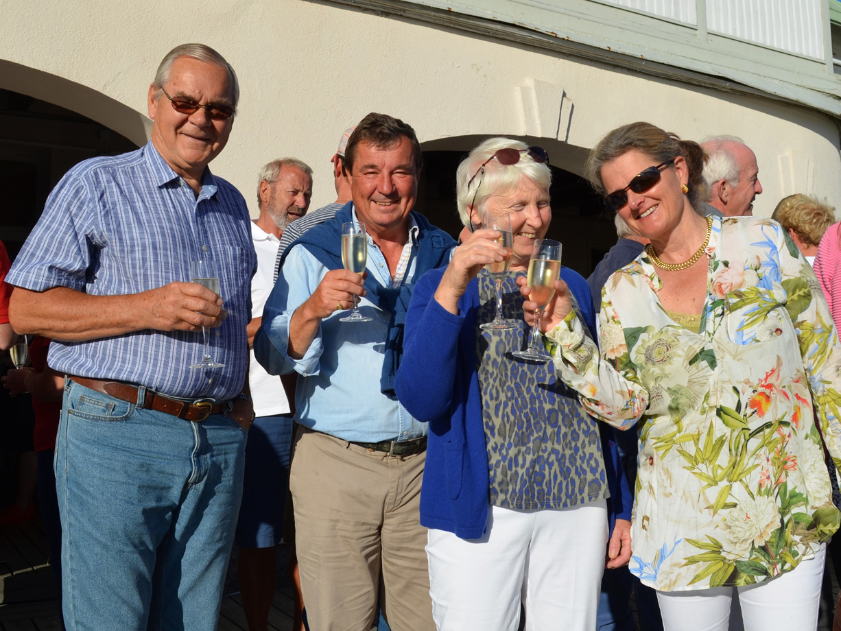 Two men and two women hold gasses of champagne and toast the camera outside a stone building on a sunny evening