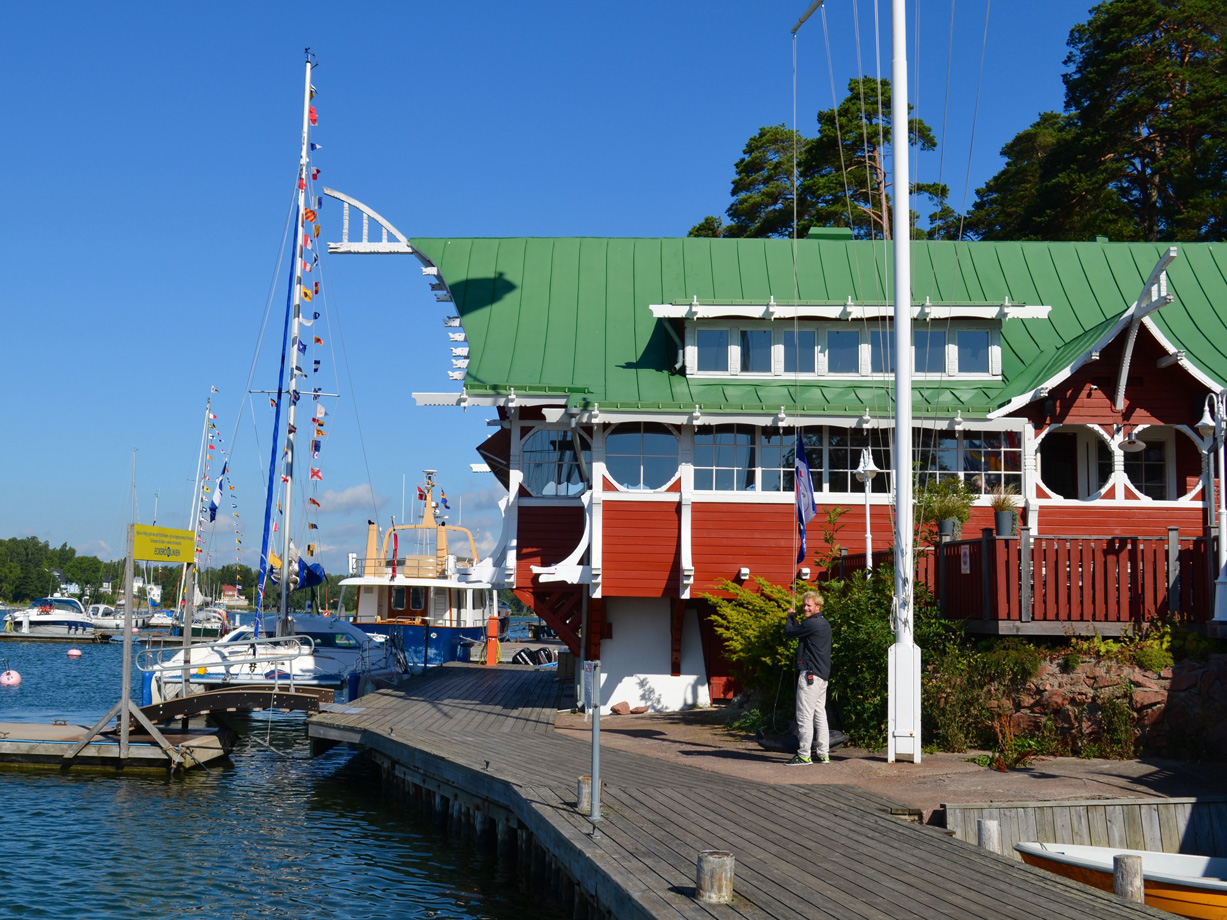 A wooden yacht dock with  boats moored alongside and a grand red wooden building with a green roof and white details