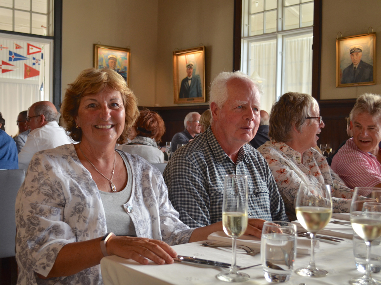 A woman smiles at the camera. She is sitting at a long table for dinner, surrounded by men and women chatting in a formal dining room