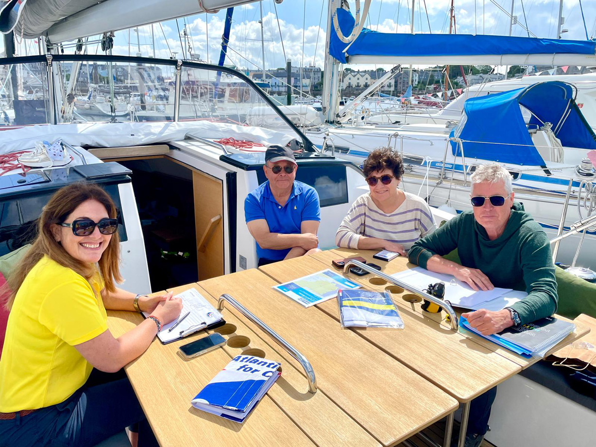 A woman wearing yellow seated at the cockpit table of a docked yacht talking to three people