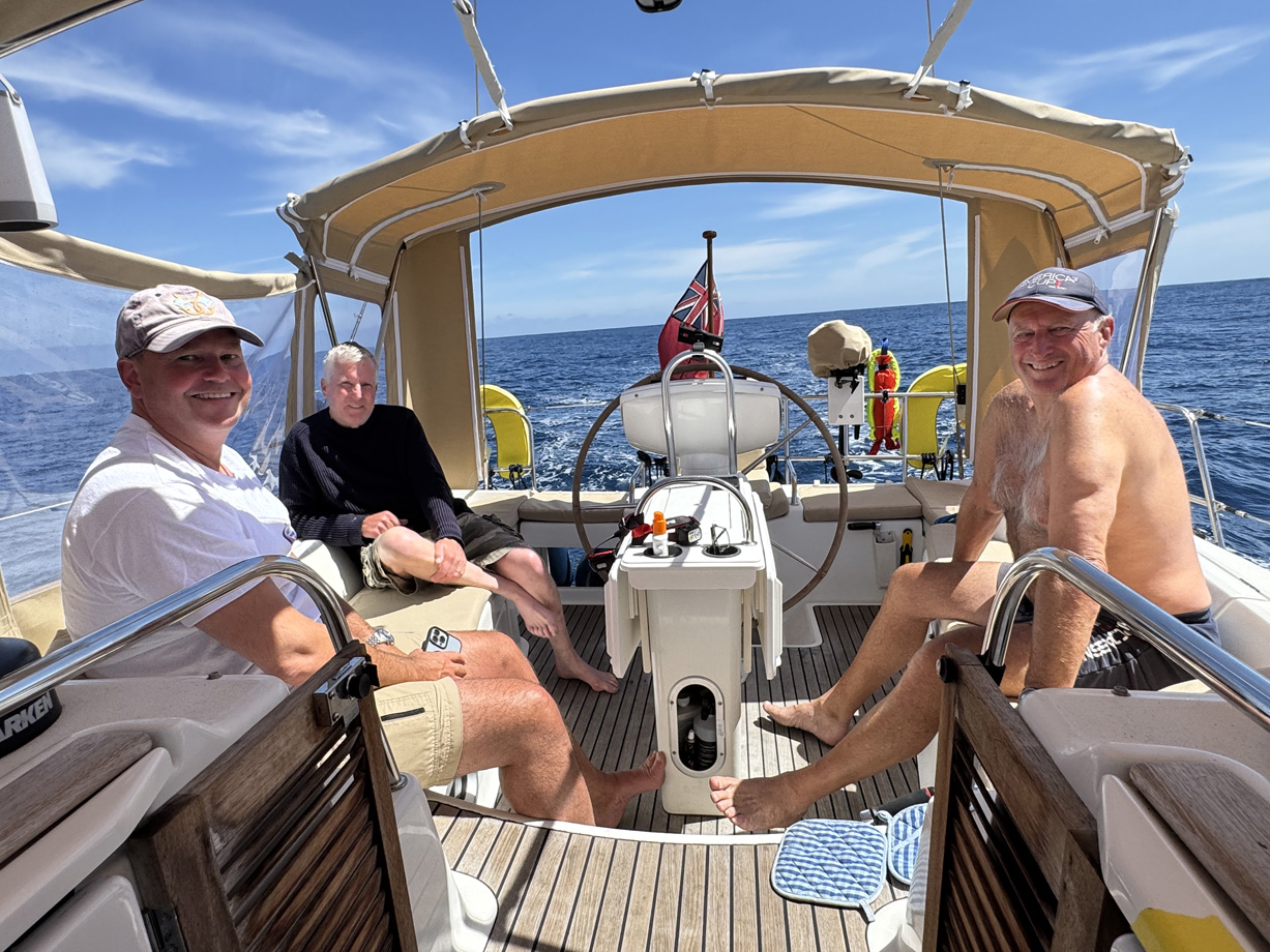 Three men site in the cockpit of a yacht at sea on a hot sunny day enjoying calm conditions
