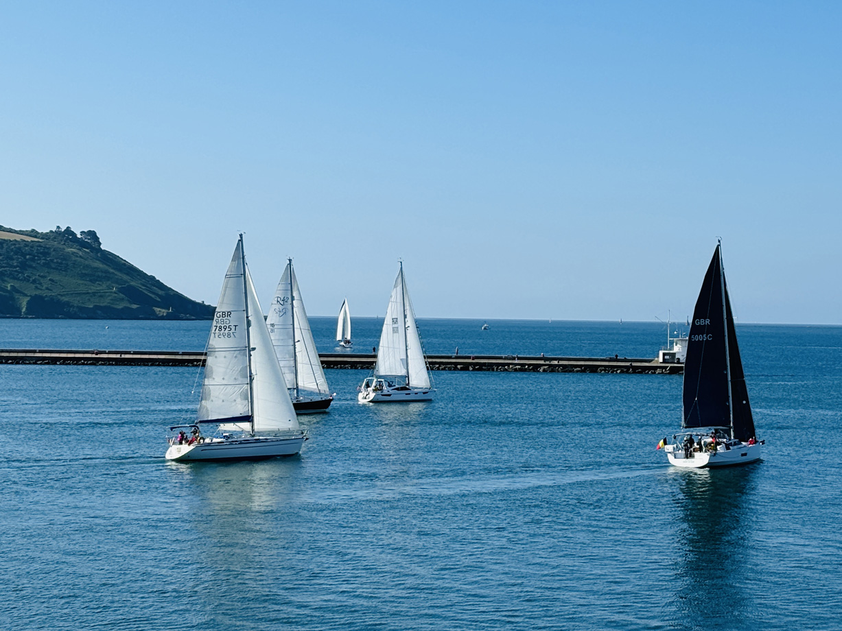 Four yachts sail towards the entrance of Plymouth breakwater with a steep green hillside in the background