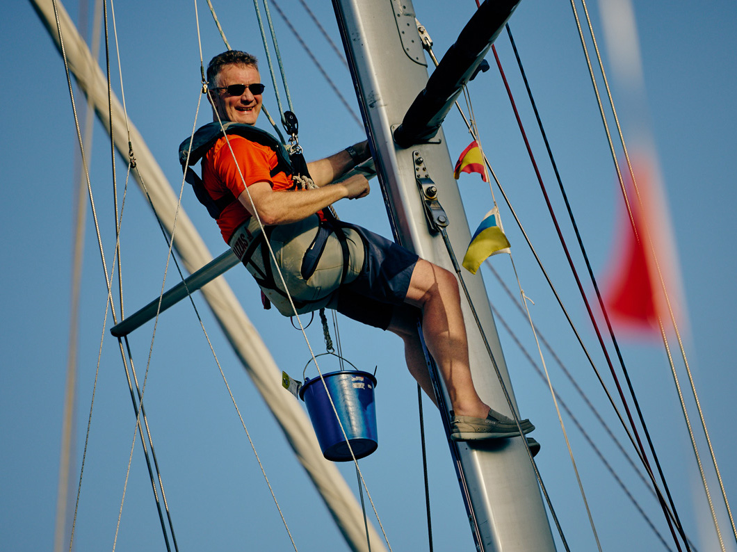 A man up the rig of a yacht in Las Palmas sits in a bosuns chair and carries his tools in a bucket