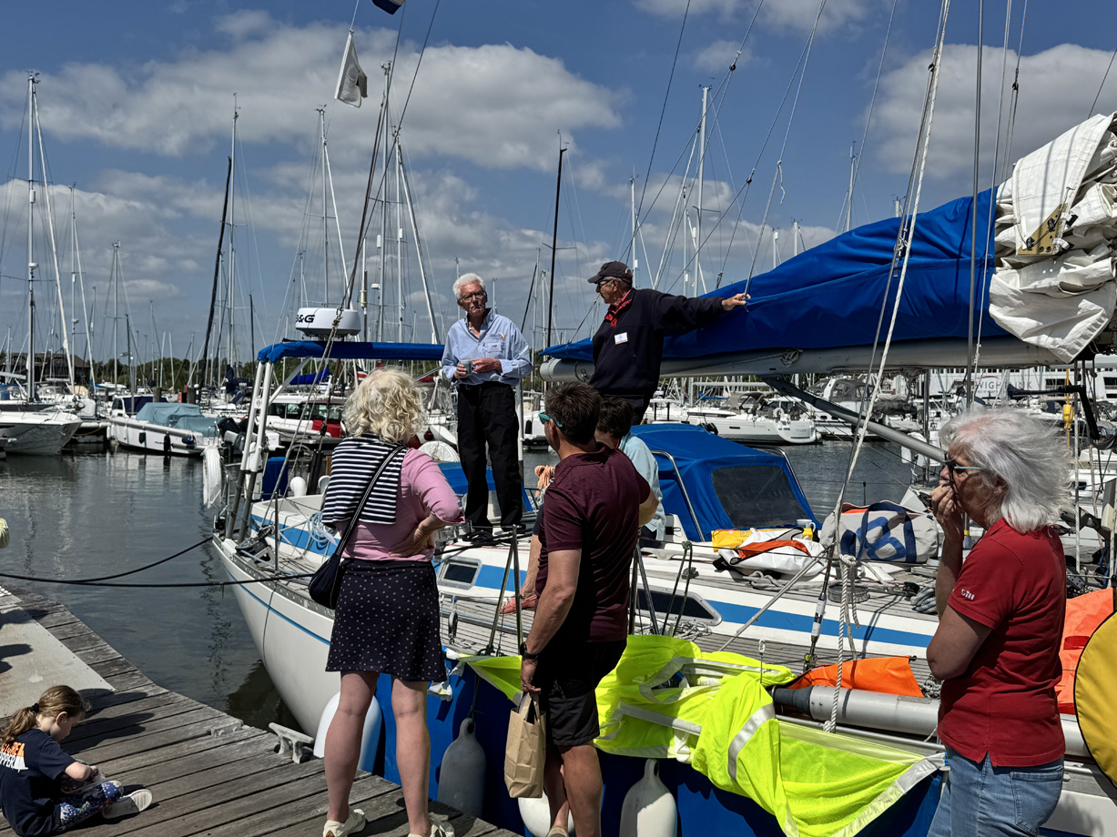 People stand on the marina dock watching a safety equipment demonstration on the deck of a yacht